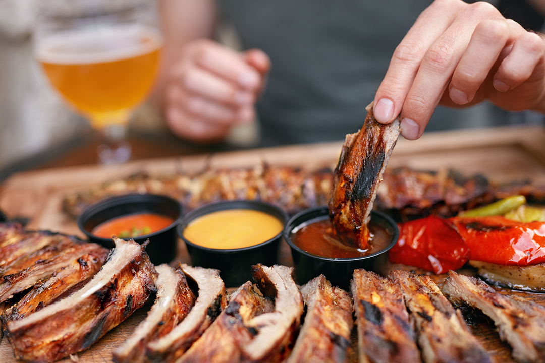 Person dipping succulent grilled ribs into a variety of BBQ sauces, with a refreshing beer in the background, highlighting a perfect barbecue feast