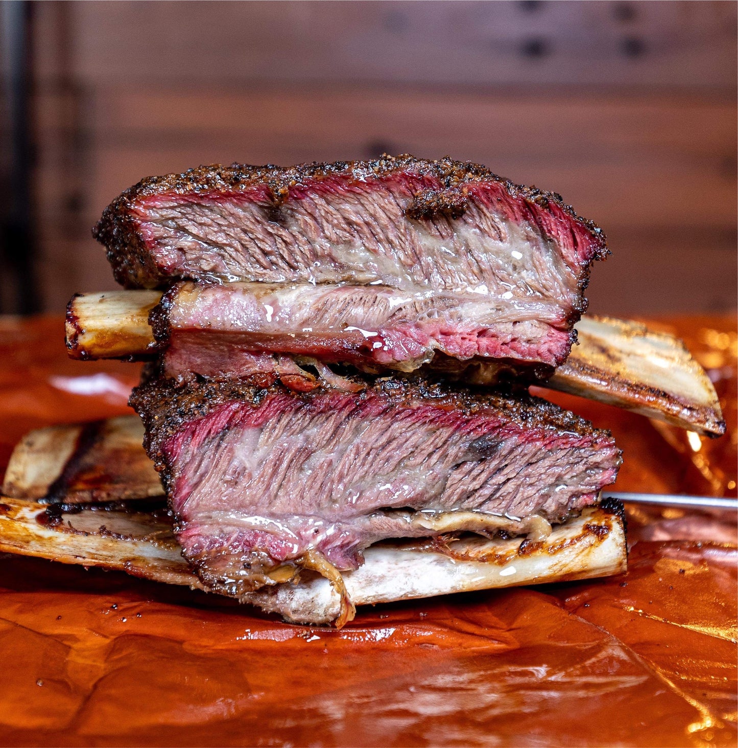 Two beef short ribs stacked on top of each other, with a dark crusty bark coating the meat. The ribs are positioned on a cutting board with a knife resting on the board next to the ribs. The meat appears juicy and tender, with visible marbling throughout.